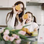 From below of cheerful mother and daughter in domestic clothes smiling and playing with oranges while sitting together at table with bouquet of tulip and drawing of child