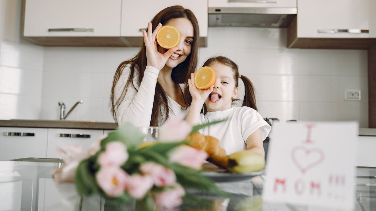 From below of cheerful mother and daughter in domestic clothes smiling and playing with oranges while sitting together at table with bouquet of tulip and drawing of child